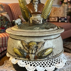 Puede incluir: Una alcancía musical de cerámica con un esmalte blanco y marrón. La alcancía presenta un busto esculpido de Johann Sebastian Bach con mazorcas de maíz a cada lado. La alcancía tiene un cordero blanco en el lateral y un querubín en la parte delantera. La base de la alcancía tiene un encaje blanco y una base negra con un interruptor de encendido/apagado.