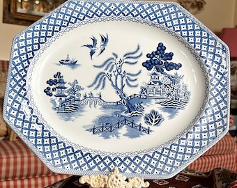 May include: A white octagonal serving platter with a blue and white willow pattern. The platter has a decorative border of blue and white diamonds. The platter is on a decorative stand with three legs.