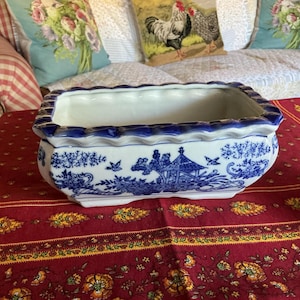 May include: A rectangular ceramic planter featuring a white base with intricate blue willow-style patterns depicting pagodas, trees, and birds. The vessel has a scalloped, wavy rim with a deep blue border and a smooth, glossy finish throughout.
