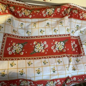 May include: A rectangular tablecloth with a red and white floral pattern. The tablecloth features a red border with white daisies and green leaves. The center has a red rectangle with a bouquet of white daisies. The background is white with yellow floral accents.