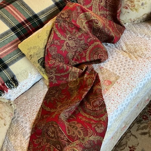 May include: A long, rectangular fabric runner featuring a deep red background with an intricate, repeating gold and brown paisley pattern. The textile has a woven texture and is draped across a surface with various patterned pillows.