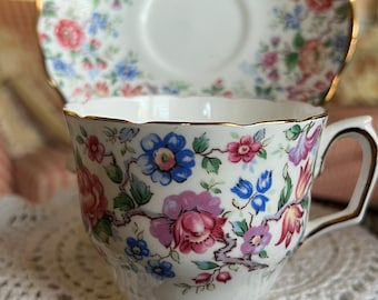 Può includere: Una tazza da tè e un piattino bianchi con un motivo floreale in tonalità di rosa, blu e verde. La tazza e il piattino hanno un bordo dorato.
