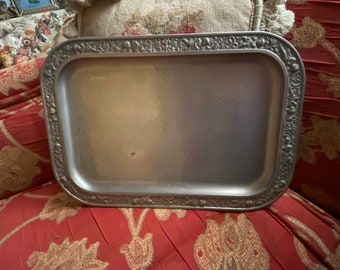 Puede incluir: Una bandeja de servir plateada con un borde decorativo en relieve. La bandeja tiene un acabado ligeramente martillado y es rectangular.