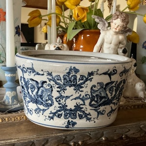 May include: An oval ceramic bowl with a cream-colored base and a blue transferware pattern featuring winged cherubs, scrolling foliage, and floral motifs. The bowl has a smooth, rounded rim and a wide, tapered body with a glossy finish.