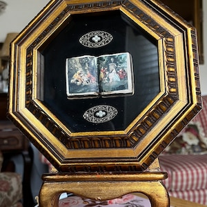 Könnte beinhalten: Ein kleines, antikes Buch mit Illustrationen wird in einem goldenen achteckigen Rahmen auf einem dekorativen Ständer ausgestellt. Das Buch ist geöffnet, um zwei Szenen zu zeigen, und es gibt zwei verzierte silberne Broschen mit Perlenakzenten über und unter dem Buch.