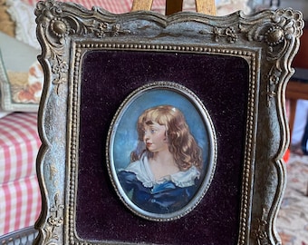 Puede incluir: Un retrato en miniatura enmarcado de una persona joven con cabello rubio que lleva un vestido azul y un cuello blanco. El marco es ornamentado y dorado con un paspartú de terciopelo morado oscuro.
