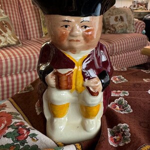 May include: A ceramic figurine of a man wearing a black hat and a burgundy jacket. He is holding a book in his hands and is sitting on a brown and white floral tablecloth.