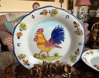 May include: A white ceramic plate with a blue rim and a hand-painted design of a rooster with a blue tail and yellow, pink, and brown feathers. The plate has a blue and white floral pattern around the rim.
