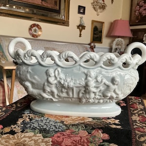 May include: A white ceramic centerpiece bowl featuring a relief design of cherubs and floral motifs. The bowl has a scalloped, openwork rim and two curved handles on either side, resting on an oval base with a smooth, glossy finish.