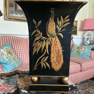 Könnte beinhalten: Schwarze dekorative Vase mit einem Pfauenmuster. Die Vase hat einen ausgestellten oberen Teil und einen rechteckigen Sockel mit goldenen Kugelfüßen. Der Pfau ist mit orangefarbenen, grünen und gelben Federn dargestellt, die auf einem belaubten Zweig sitzen.