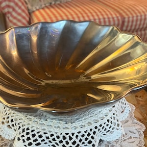 May include: A silver, scallop-shaped bowl with a shiny, reflective surface. The bowl is sitting on a white crocheted doily.