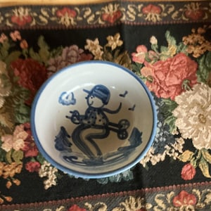May include: A small, round, white ceramic bowl with a blue rim. The interior features a hand-painted blue illustration of a person wearing a hat and scarf, skiing on a snowy surface with stylized trees in the background and a small cloud above.