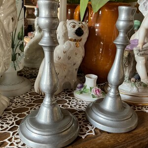 May include: Two antique silver-colored candlesticks with a classic design. The candlesticks have a wide base and a slender, curved stem. The background includes decorative ceramic figurines and a vase.