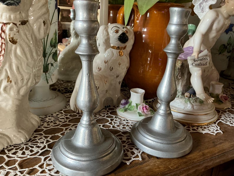 May include: Two antique silver-colored candlesticks with a classic design. The candlesticks have a wide base and a slender, curved stem. The background includes decorative ceramic figurines and a vase.