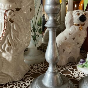 May include: A silver-colored candlestick with a round base and a slender, detailed stem. The candlestick is placed on a lace doily, with white ceramic dog figurines and other decorative items in the background.