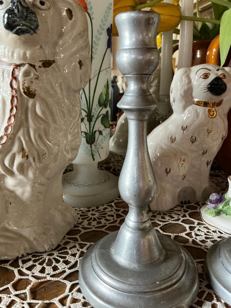 May include: A silver-colored candlestick with a round base and a slender, detailed stem. The candlestick is placed on a lace doily, with white ceramic dog figurines and other decorative items in the background.