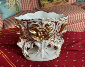 Puede incluir: Jarrón de cerámica blanca con detalles dorados. El jarrón presenta un borde festoneado y diseños florales y de hojas doradas. La base tiene forma redondeada con detalles calados. Una pieza decorativa para el hogar.