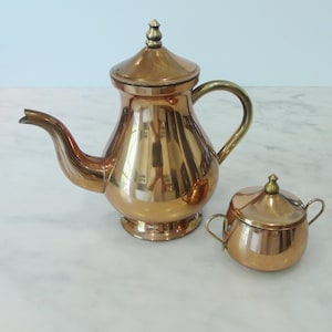 May include: A copper teapot and sugar bowl set with brass accents. The teapot has a rounded body and a lid with a finial. The sugar bowl has a round body with two handles and a lid with a finial.