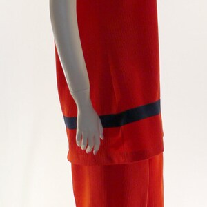 May include: A red ribbed jumpsuit with a black trim around the neckline and sleeves. The jumpsuit has a short-sleeved top and wide-legged pants.