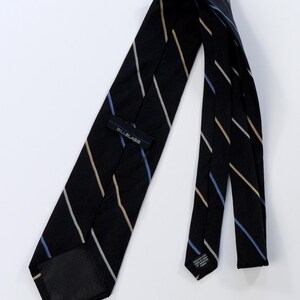 Puede incluir: Una corbata negra con rayas diagonales blancas, beige y azules. La etiqueta dice "BILLBLASS". La corbata es de seda, un accesorio cl&aacute;sico.