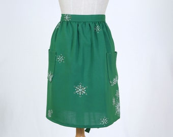 May include: A green apron with white snowflake designs. The apron has two pockets and a tie at the waist.