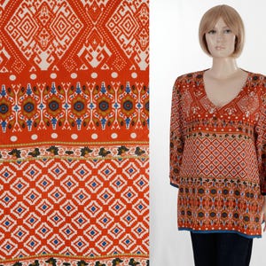May include: A long-sleeve tunic featuring a vibrant orange base with a multi-patterned design. The garment displays horizontal bands of geometric shapes, diamonds, and floral motifs in white, blue, and dark brown, finished with a V-neckline.