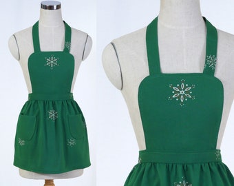 May include: A green apron with white and brown snowflake embroidery. The apron has a bib top with shoulder straps, a gathered skirt, and two front pockets. The apron is displayed on a mannequin.