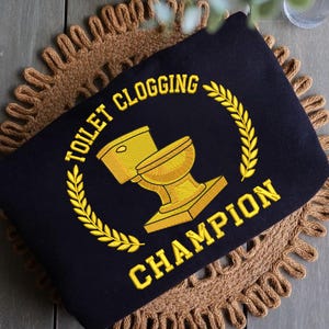 May include: Black rectangular pillow with gold embroidered text and a toilet graphic. The text reads "TOILET CLOGGING CHAMPION" around a gold toilet graphic, encircled by a gold laurel wreath.