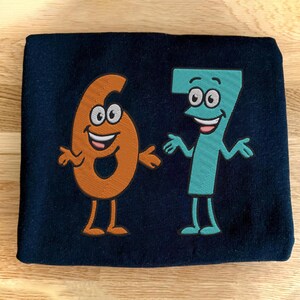 K&ouml;nnte beinhalten: Marineblauer Stoff mit gestickten Cartoon-Zahlen sechs und sieben. Die Zahl sechs ist orange und die Zahl sieben ist t&uuml;rkis. Beide Zahlen haben l&auml;chelnde Gesichter, Arme und Beine.