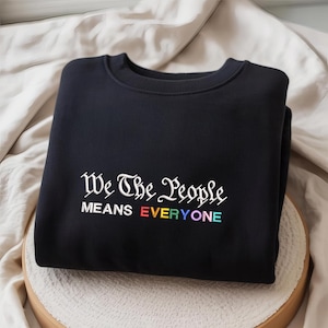 May include: Black sweatshirt with the text "We The People MEANS EVERYONE" in white and rainbow colors. The sweatshirt is folded and resting on a light-colored surface.