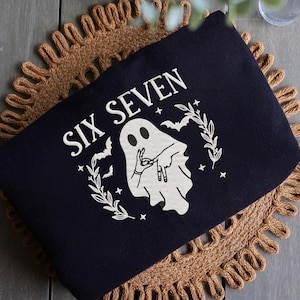 May include: A dark blue pouch with the words "SIX SEVEN" in white, along with a white ghost illustration making a hand gesture. The design includes bats, stars, and a leafy wreath. The pouch is on a brown woven mat.