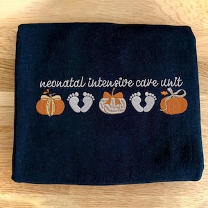Pode incluir: Cobertor azul marinho bordado com as palavras "neonatal intensive care unit". O design inclui duas abóboras laranja, dois pares de pegadas de bebê brancas e uma abóbora com um laço.