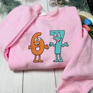 K&ouml;nnte beinhalten: Hellrosa Sweatshirt mit den gestickten Zahlen 6 und 7 als Cartoonfiguren. Die Zahl 6 ist orange und die Zahl 7 ist t&uuml;rkis. Beide Zahlen haben l&auml;chelnde Gesichter und Arme.