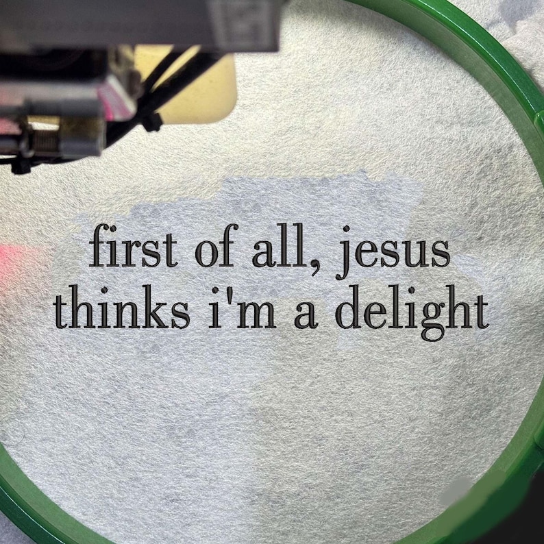Pu&ograve; includere: Primo piano di tessuto bianco con la scritta "first of all, jesus thinks i'm a delight" in nero. Un telaio da ricamo verde incornicia il tessuto. L'immagine mostra anche una parte di una macchina da ricamo.