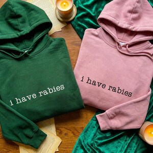 Könnte beinhalten: Zwei Kapuzenpullover, einer in Waldgrün und einer in Staubrosa, werden gezeigt. Jeder Pullover hat den weißen Aufdruck "i have rabies". Sie liegen auf einer Holzoberfläche mit einer Kerze und Samtstoff.
