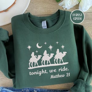 May include: Forest green sweatshirt with a white graphic of three figures riding camels under a crescent moon and stars. Text reads "tonight, we ride. Matthew 2:1". The sweatshirt is folded on a wooden surface.