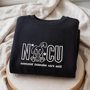 May include: Black sweatshirt with white text and a bunny graphic. The text reads "NICU" with a bunny holding an egg and the words "neonatal intensive care unit" below. The sweatshirt is folded on a light-colored surface.