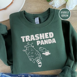May include: Forest green sweatshirt featuring the text "Trashed Panda" and a graphic of a raccoon and spilled bottle. The sweatshirt is folded, with a white hat and flower in the background. The sweatshirt is made of a soft material.