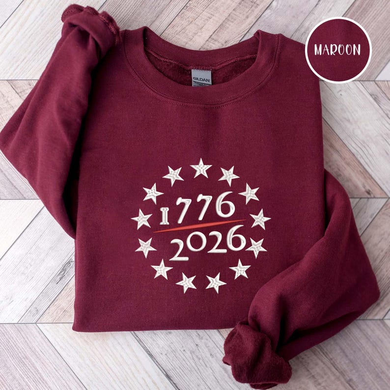 May include: Maroon sweatshirt with a white star-studded circle design. The design includes the numbers "1776" above a horizontal line and "2026" below. The word "MAROON" is in a circle in the upper right corner.