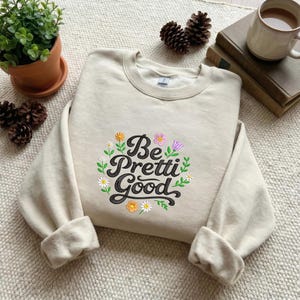 Pode incluir: Camisola bege com a frase bordada "Be Pretti Good" rodeada por flores coloridas e folhas verdes. A parte superior de manga comprida está dobrada sobre uma superfície texturizada.