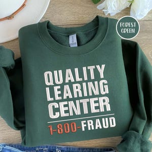 Könnte beinhalten: Ein dunkelgrünes Sweatshirt mit dem weißen Schriftzug „QUALITY LEARNING CENTER“ und der orangefarbenen Aufschrift „1-800-FRAUD“. Das Sweatshirt ist gefaltet, im Hintergrund sind ein weißer Hut und blaue Jeans zu sehen.