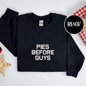 May include: Black crewneck sweatshirt with the text "PIES BEFORE GUYS" in white block letters. A black circle with the word "BLACK" is in the upper right corner. The sweatshirt is on a white surface.