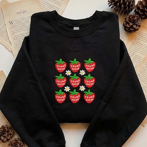 May include: Black crewneck sweatshirt featuring an embroidered design of strawberries with the text "TRUMP SUCKS" in red. White daisy accents are interspersed between the strawberries. The word "twilight" is visible in the background.
