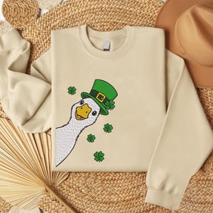 May include: A cream-colored sweatshirt featuring a cartoon goose wearing a green St. Patrick's Day hat with a gold buckle and shamrocks. The goose is peeking from the left side, surrounded by green shamrock graphics. The sweatshirt has long sleeves.