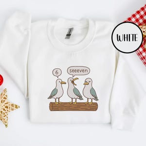 May include: White sweatshirt with a graphic of three cartoon seagulls on a log. One seagull has a thought bubble with the number 6, and another has a speech bubble that says "seeeven". The word "WHITE" is in a black circle.