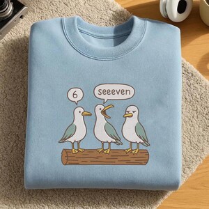 May include: Light blue sweatshirt featuring a humorous illustration of three seagulls perched on a log. One seagull says "6" and another says "seeeven". The seagulls are white with gray and teal accents. The sweatshirt has a crew neck.