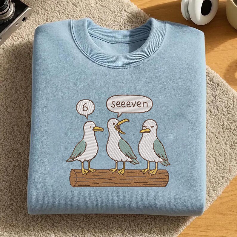 May include: Light blue sweatshirt featuring a humorous illustration of three seagulls perched on a log. One seagull says "6" and another says "seeeven". The seagulls are white with gray and teal accents. The sweatshirt has a crew neck.