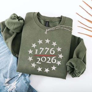 May include: An olive green sweatshirt with the numbers "1776" and "2026" in white, surrounded by a circle of stars. A silver necklace with a small pendant rests on the sweatshirt. The sweatshirt is folded over a pair of blue jeans.