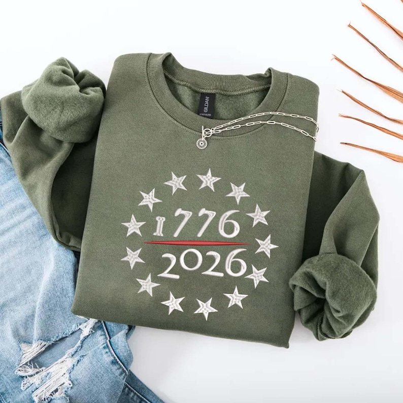 May include: An olive green sweatshirt with the numbers "1776" and "2026" in white, surrounded by a circle of stars. A silver necklace with a small pendant rests on the sweatshirt. The sweatshirt is folded over a pair of blue jeans.