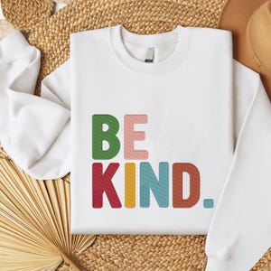 May include: White sweatshirt with the embroidered text "BE KIND" in colorful letters. The letters are green, pink, red, yellow, blue, and brown. The sweatshirt has a crew neck and long sleeves.